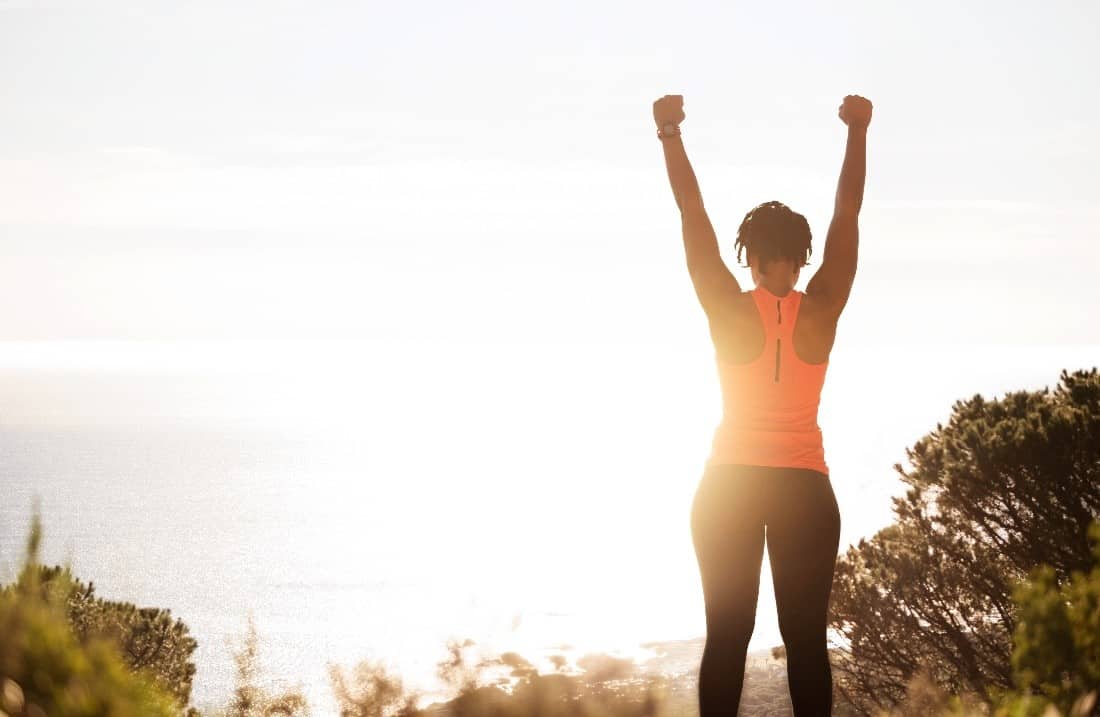 A woman in active wear raises her arms triumphantly as she completes climbing a seaside hill