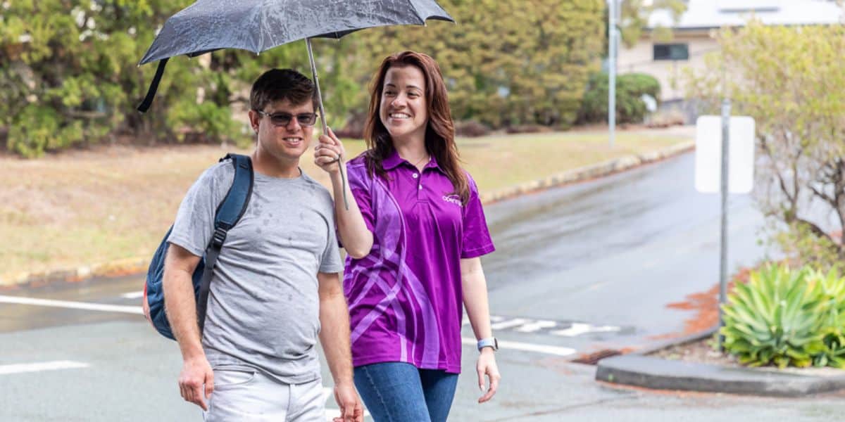 A mental health support worker holding an umbrella over a client outside in the rain.